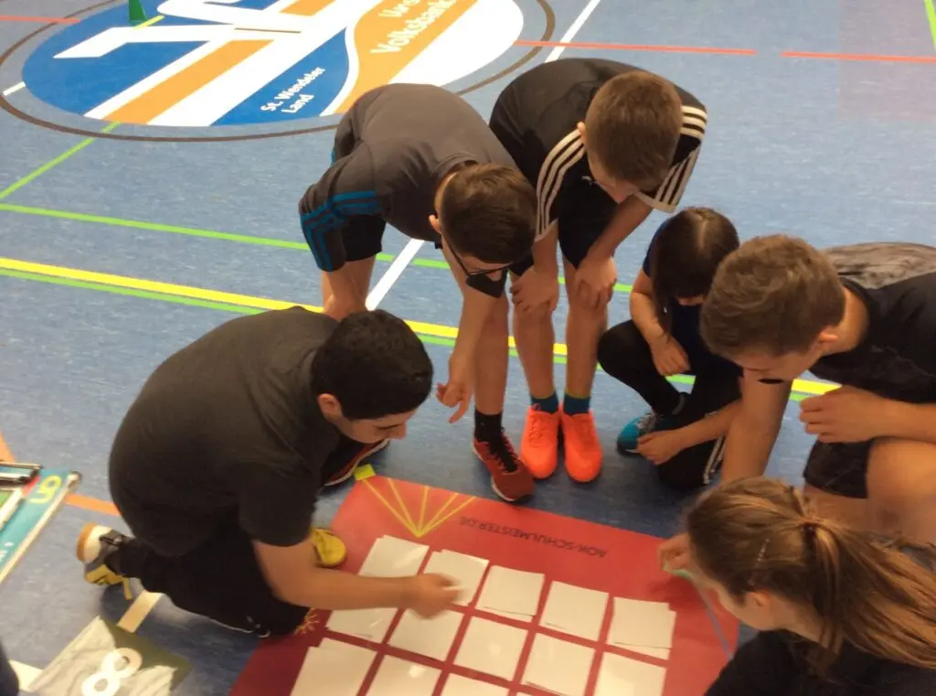Jugendliche knien um ein Spielfeld aus Klebeband auf dem Hallenboden und lösen gemeinsam eine Aufgabe mit Ball