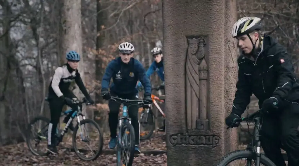 Drei Mountainbiker mit Helm fahren durch einen winterlichen Wald, rechts ein steinernes Wegkreuz