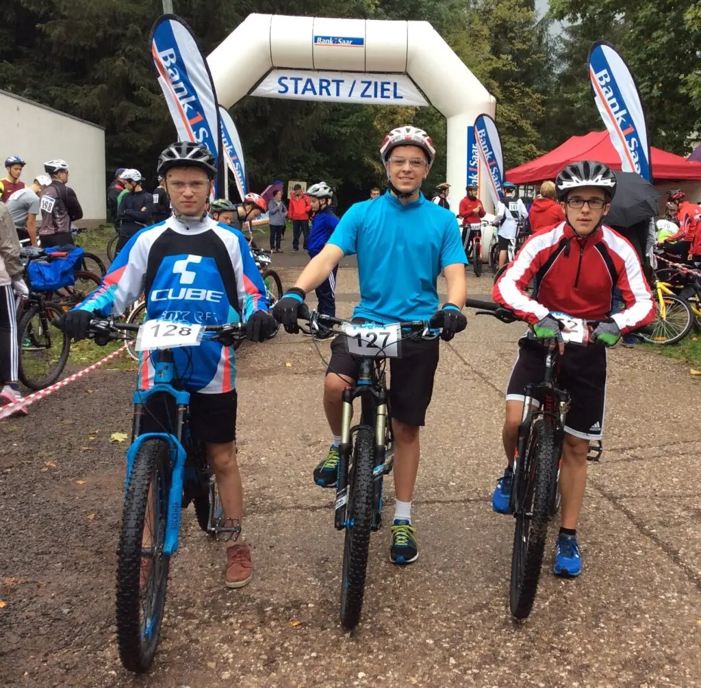 Drei Jugendliche mit Helmen auf Mountainbikes vor einem Start/Ziel-Bogen