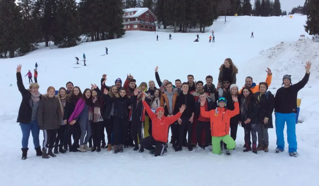 Große Gruppe Jugendlicher in Winterkleidung jubelt mit erhobenen Armen auf einer Skipiste