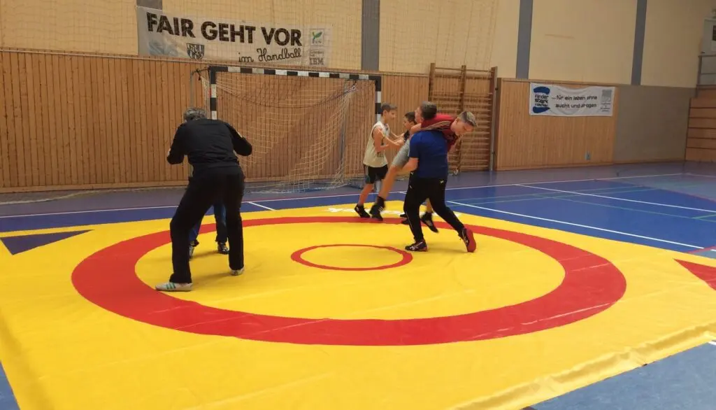 Zwei Kinder ringen auf einer gelb-roten Matte, ein Kampfrichter steht daneben, an der Wand das Banner „Fair geht vor“