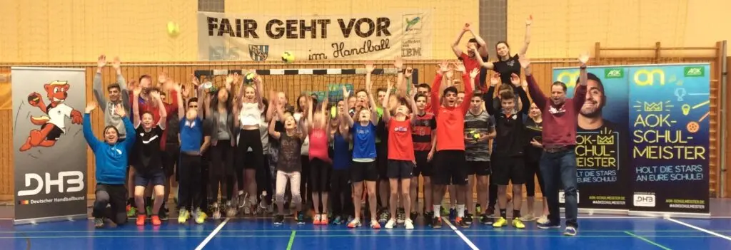 Große Schülergruppe jubelt mit erhobenen Armen in der Sporthalle unter dem Banner „Fair geht vor“, daneben AOK- und DHB-Aufsteller