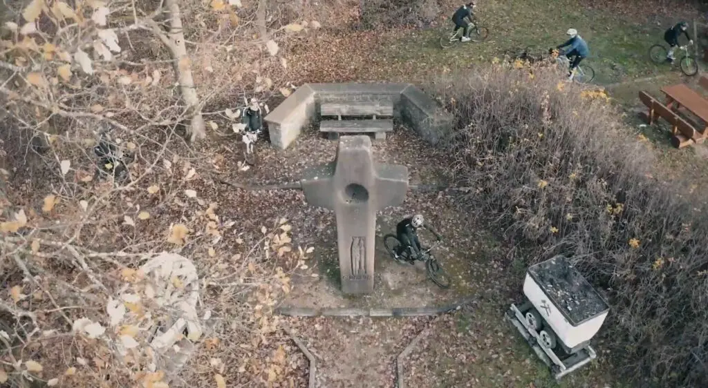 Luftaufnahme eines Waldstücks mit Gestrüpp, einem steinernen Kreuz und Holzaufbauten, am Rand einzelne Personen