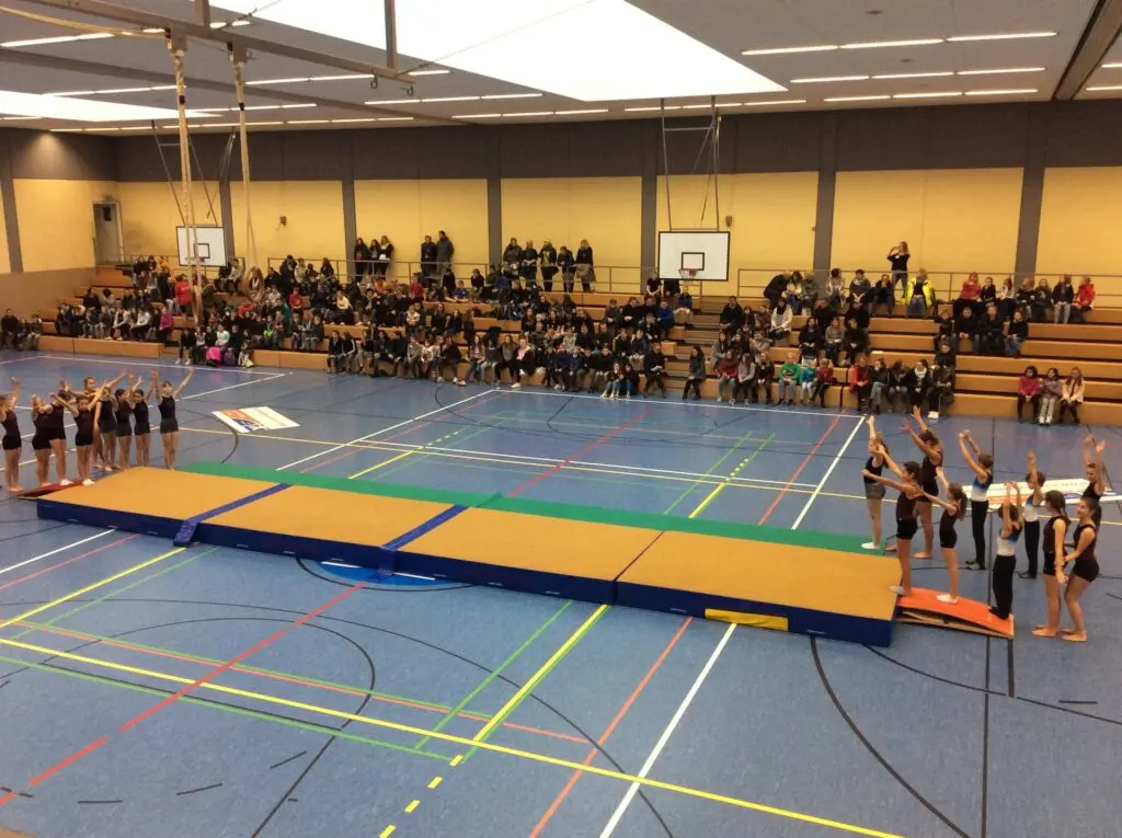 Turnstationen mit langen Mattenbahnen in einer Sporthalle, Kindergruppen und Zuschauer auf der Tribüne