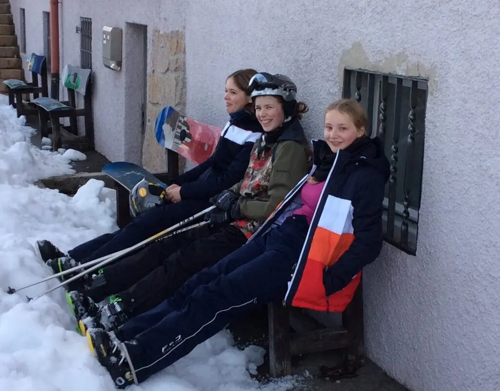 Jugendliche in Skikleidung sitzen auf einer Bank vor einer Hüttenwand im Schnee