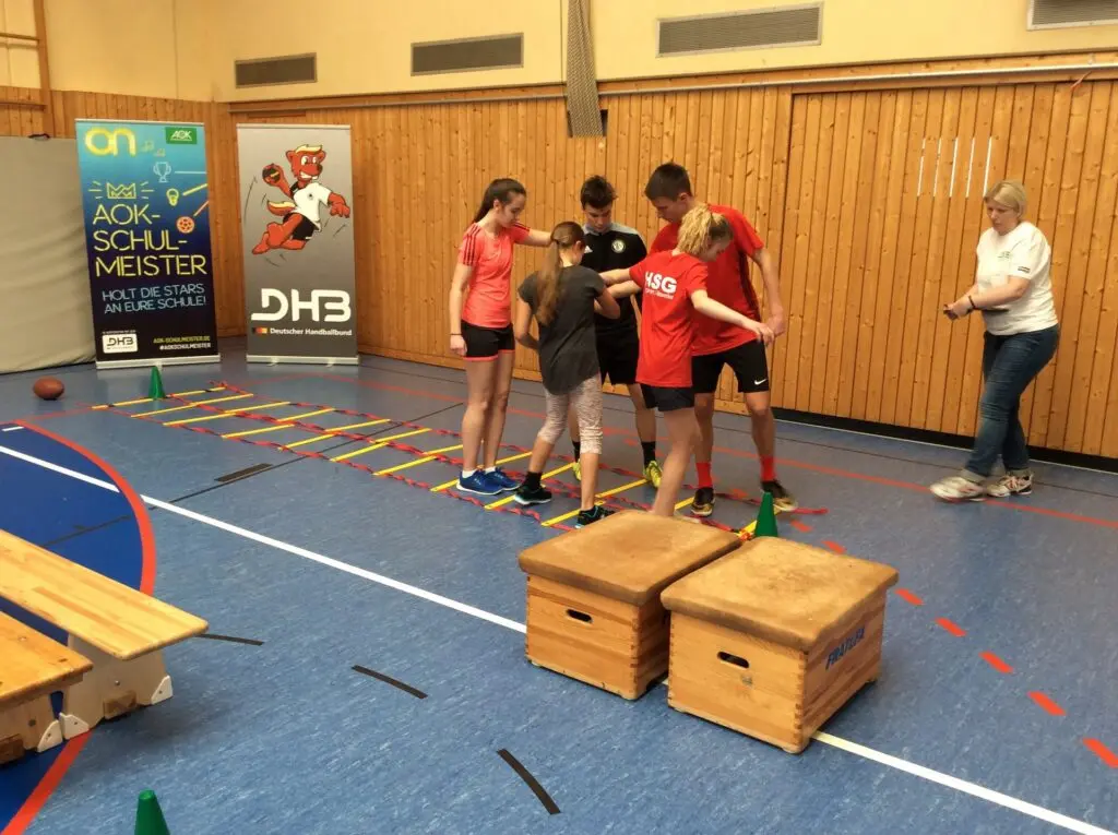 Schülergruppe bewältigt einen Hindernisparcours mit Holzkästen und Koordinationsleiter in der Sporthalle, eine Betreuerin schaut zu
