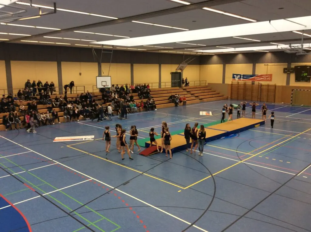 Kinder turnen auf Mattenbahnen in einer großen Sporthalle, am Rand sitzen Zuschauer