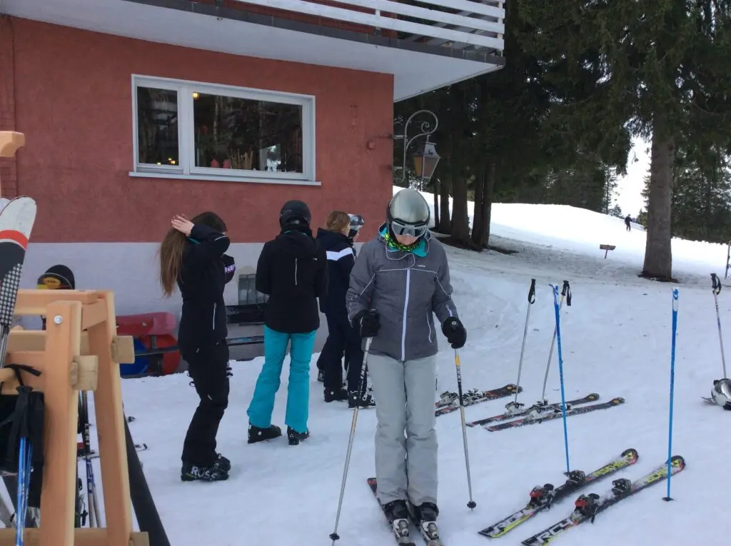 Jugendliche mit Skiausrüstung stehen vor einer roten Hütte im Schnee, Ski stecken im Schnee
