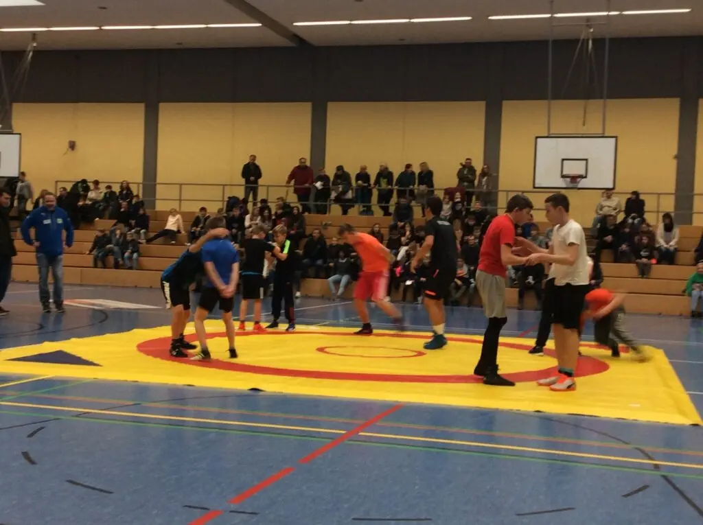 Wettkampfbetrieb auf einer großen Ringermatte in der Sporthalle, Zuschauer am Rand