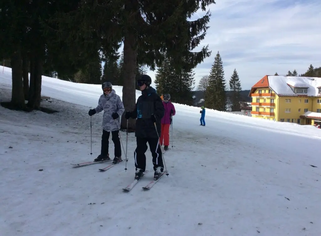 Jugendliche mit Ski stehen auf einer verschneiten Piste, im Hintergrund ein Gebäude und Tannen