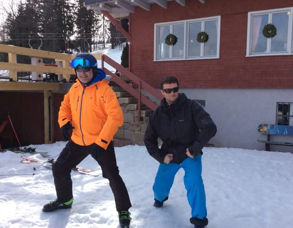 Zwei Jugendliche in Skikleidung posieren lässig im Schnee vor einer roten Hütte