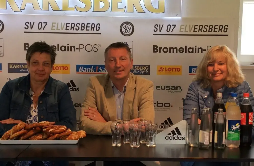 Drei Personen sitzen vor einer Sponsorenwand des SV 07 Elversberg an einem Tisch mit Getränken und Brezeln