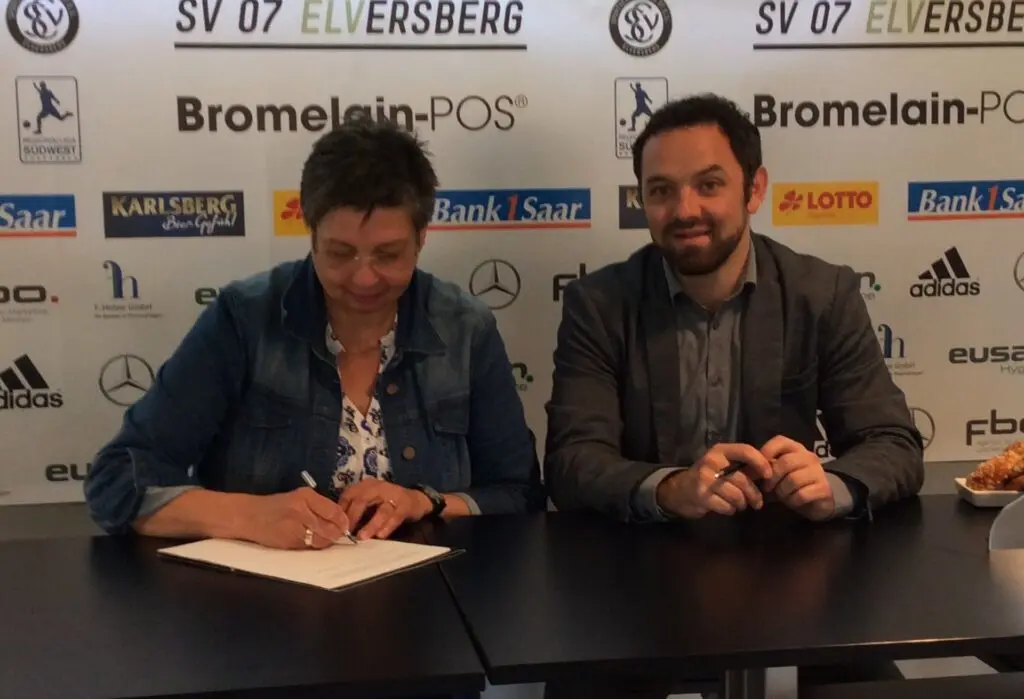 Frau unterschreibt neben einem Mann vor der Sponsorenwand des SV 07 Elversberg ein Dokument