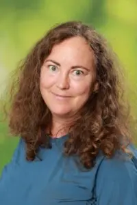 Porträt Martina Eckert, mit lockigen Haaren im blauen Oberteil vor grünem Hintergrund