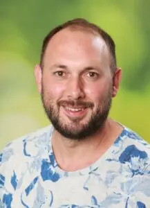 Porträt Raphael Fuchs, mit Bart im gemusterten Hemd vor grünem Hintergrund