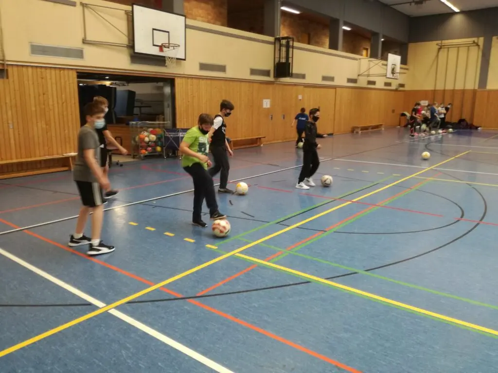 Kinder üben in der Fußball-AG Ballführung um Hütchen in einer Sporthalle