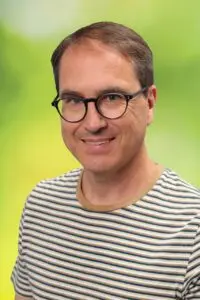 Porträt Christof Georg, mit Brille im gestreiften Pullover vor grünem Hintergrund