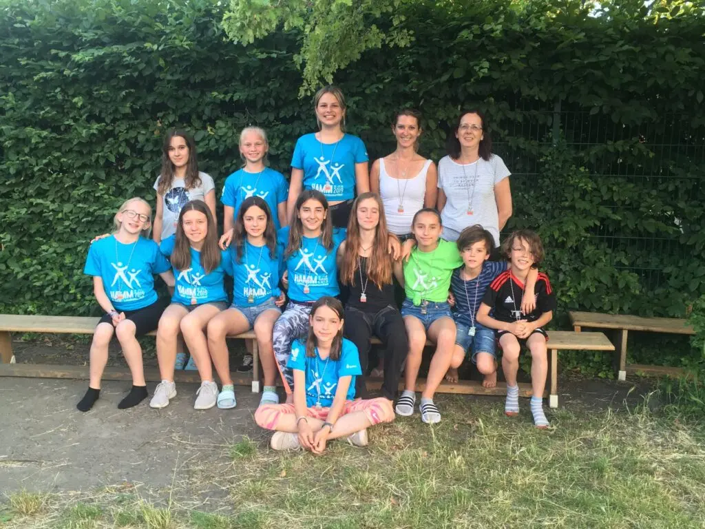 Geräteturnerinnen in blauen T-Shirts sitzen und stehen als Gruppe auf und hinter einer Holzbank vor einer Hecke