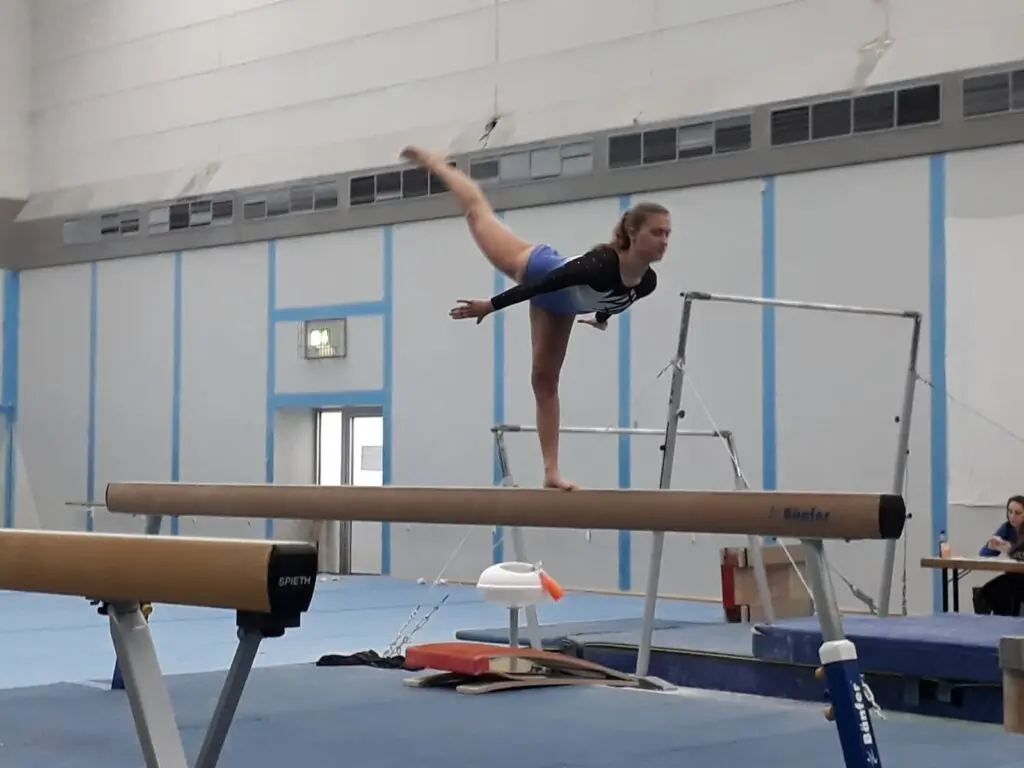 Turnerin zeigt eine Standwaage auf dem Schwebebalken in einer Sporthalle, daneben ein Stufenbarren