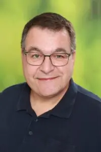 Porträt Patrick Gotthard, mit Brille im dunklen Poloshirt vor grünem Hintergrund