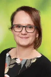 Porträt Eva Hartmann, mit Brille im dunklen Oberteil vor grünem Hintergrund