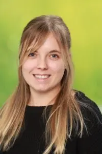 Porträt Justine Hautz, mit langen blonden Haaren im schwarzen Oberteil vor grünem Hintergrund