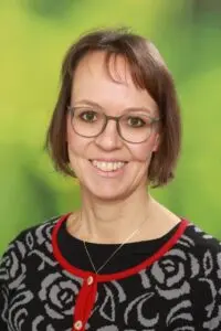 Porträt Silvia Hinsberger, mit Brille und rot-schwarzem Pullover vor grünem Hintergrund