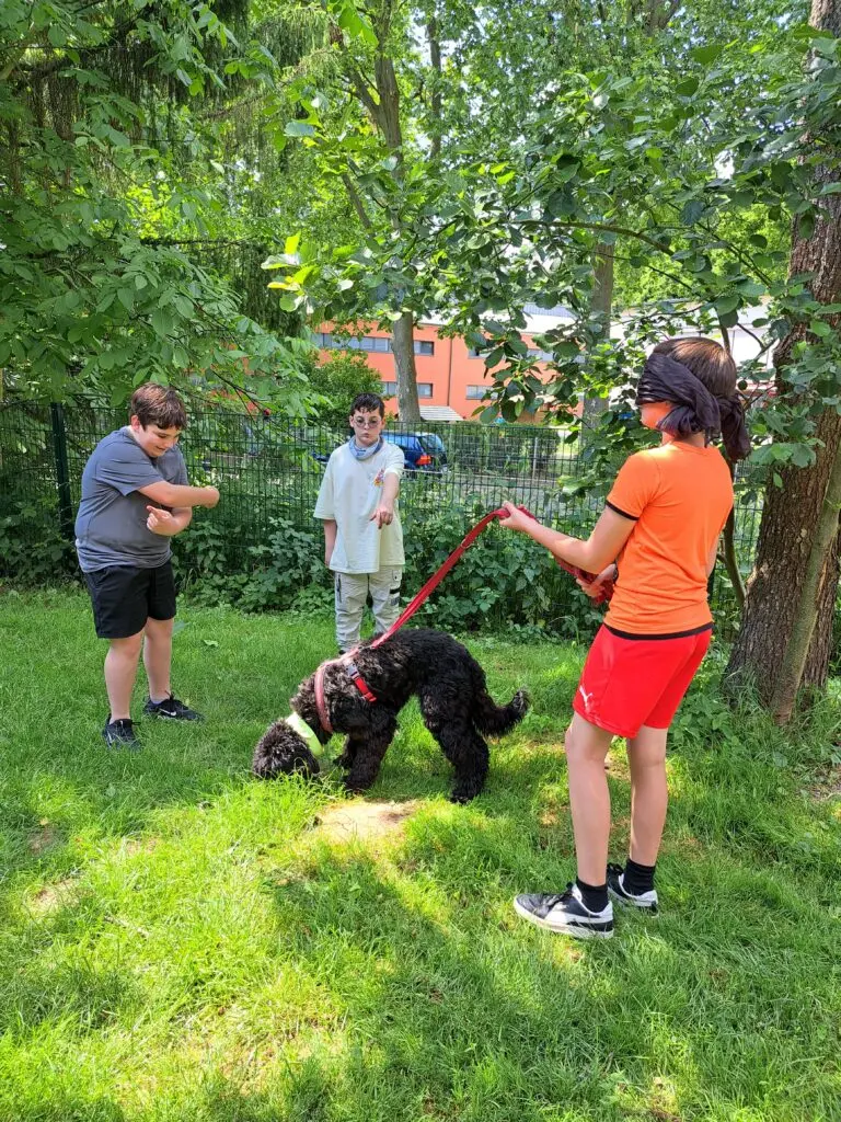 Kinder stehen auf einer Wiese im Grünen, ein Junge führt einen schwarzen Hund an der Leine
