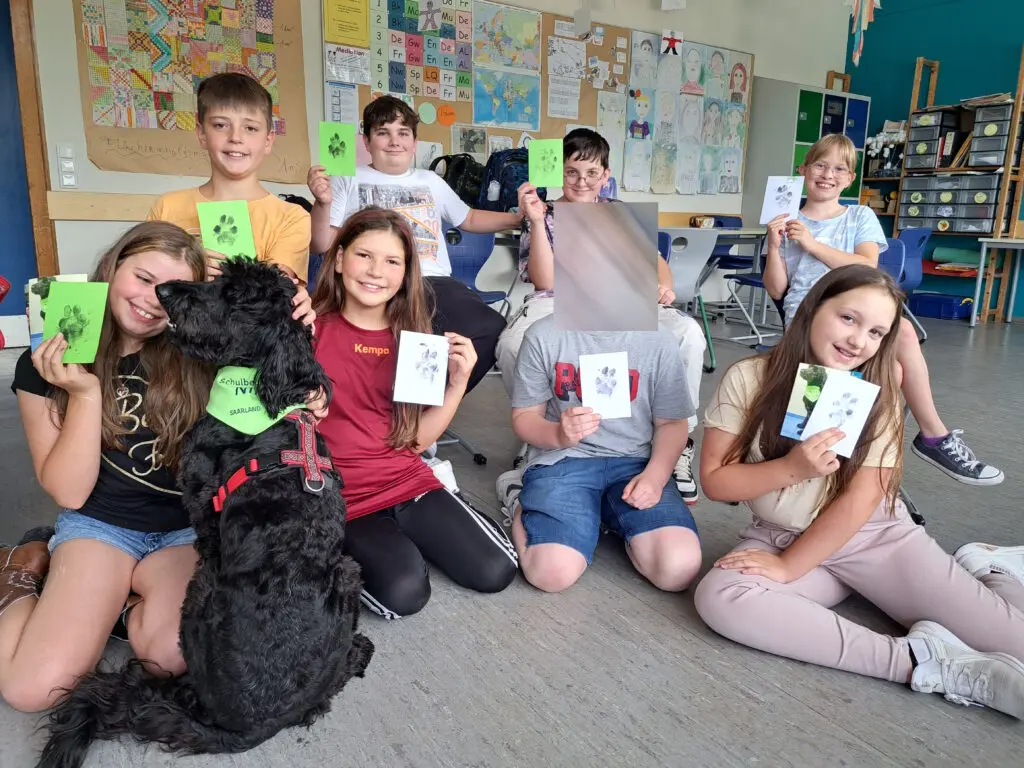 Schülerinnen und Schüler sitzen mit Karten in der Hand im Klassenraum auf dem Boden, vorne ein schwarzer Hund