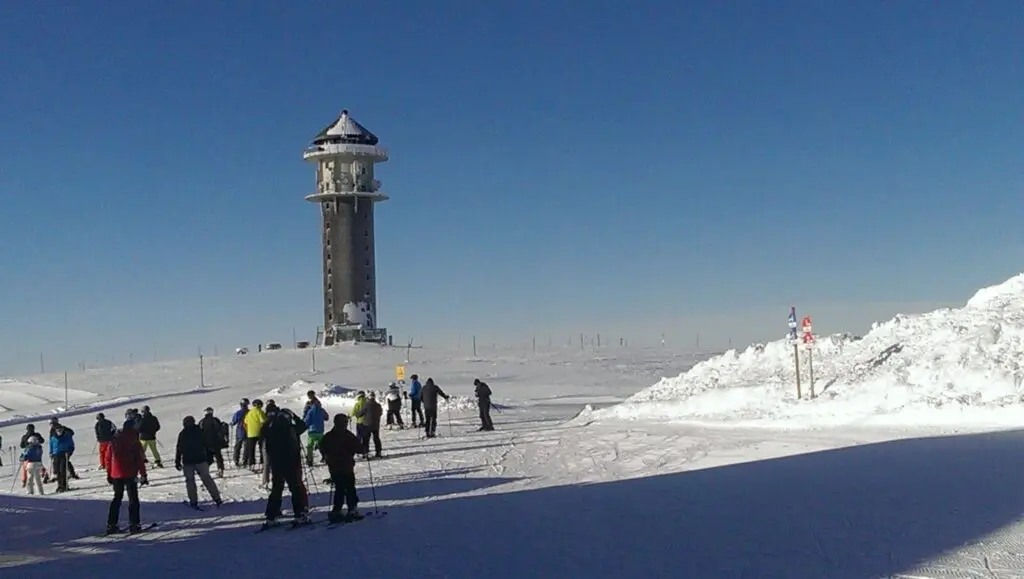 Skifahrer auf einer weiten Schneefläche unter blauem Himmel, im Hintergrund ein steinerner Aussichtsturm