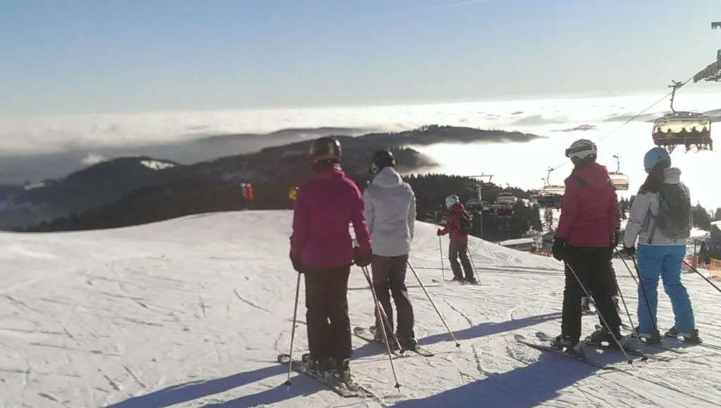 Skifahrer stehen auf einer sonnigen Piste mit Blick über ein Wolkenmeer, rechts ein Sessellift
