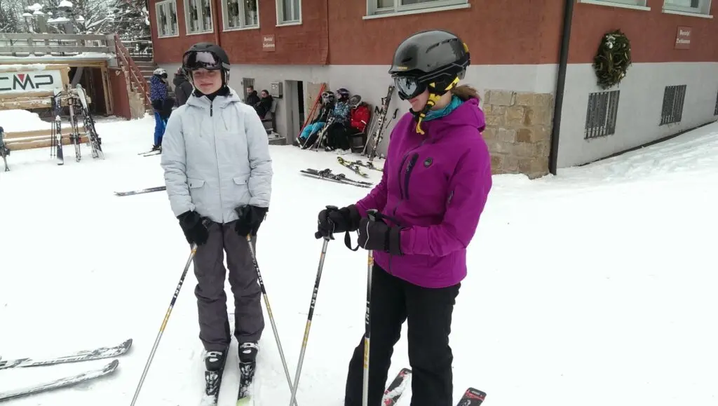 Zwei Jugendliche mit Helm und Skistöcken stehen auf der Piste vor einer Hütte