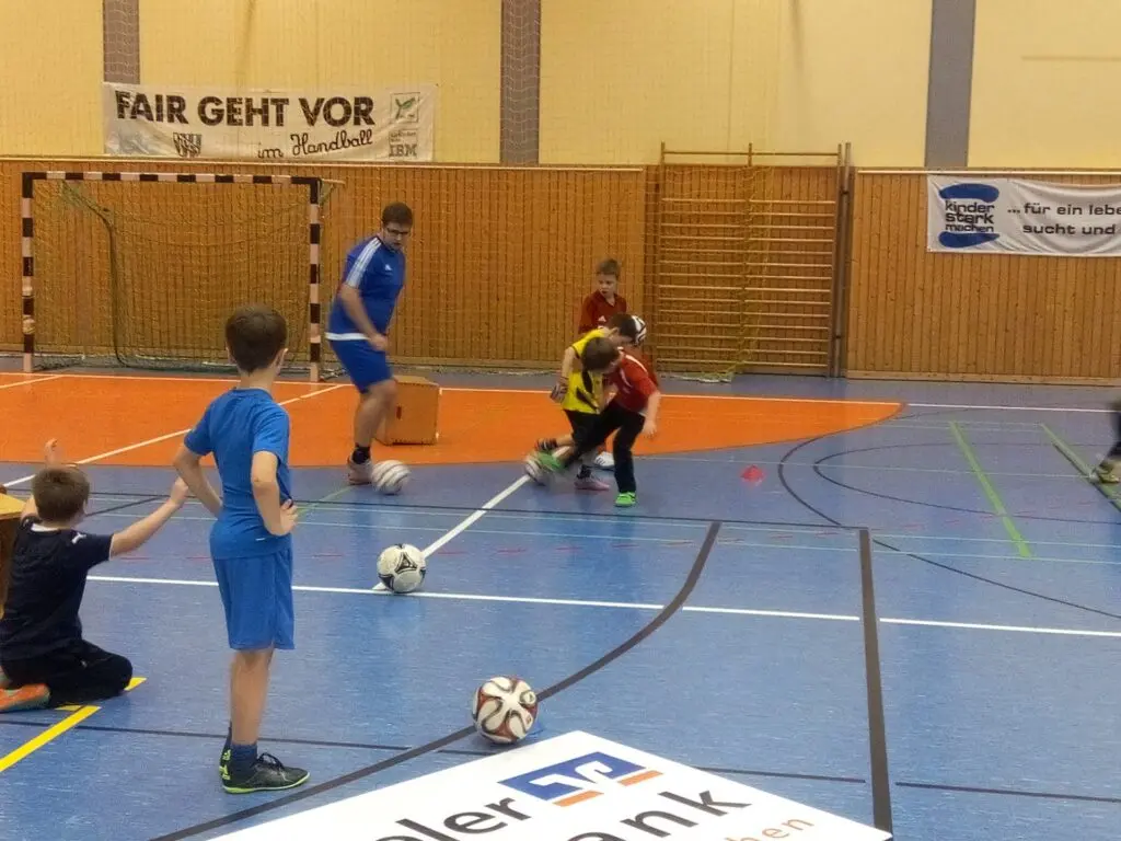 Kinder dribbeln Bälle in der Sporthalle, ein Jugendlicher betreut die Übung