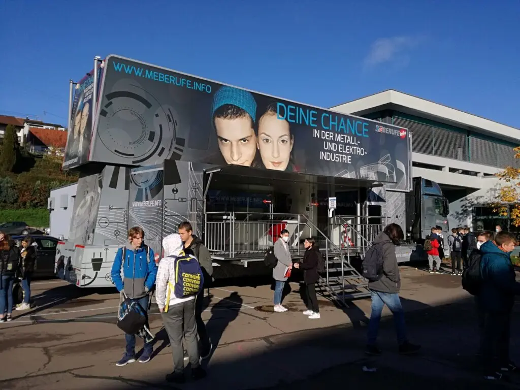 Großer ausgeklappter Infotruck auf dem Schulgelände, davor Schülerinnen und Schüler in der Sonne