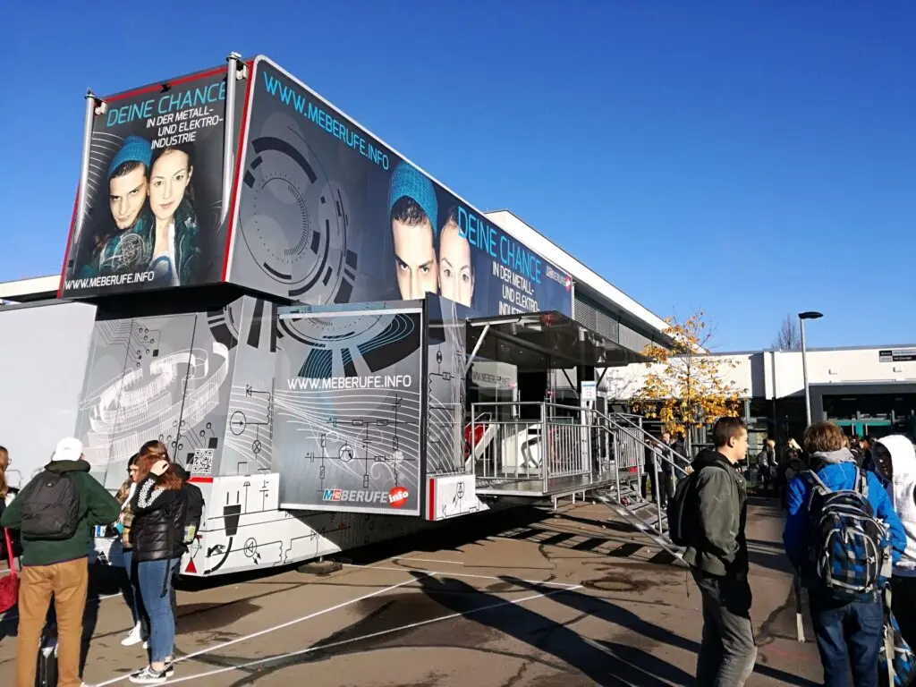 Zweistöckiger Ausstellungstruck mit Werbebildern auf einem Platz, davor Schülergruppen