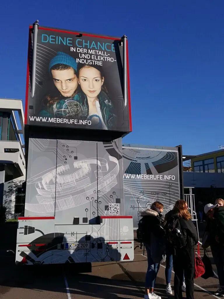 Doppelstöckiger Infotruck mit Werbeplakat vor blauem Himmel, davor Jugendliche