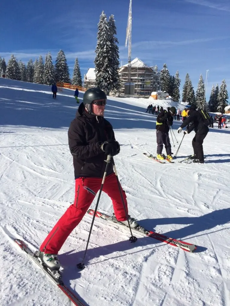Skifahrerin mit roter Hose fährt auf einer Piste, im Hintergrund Tannen und ein Turm