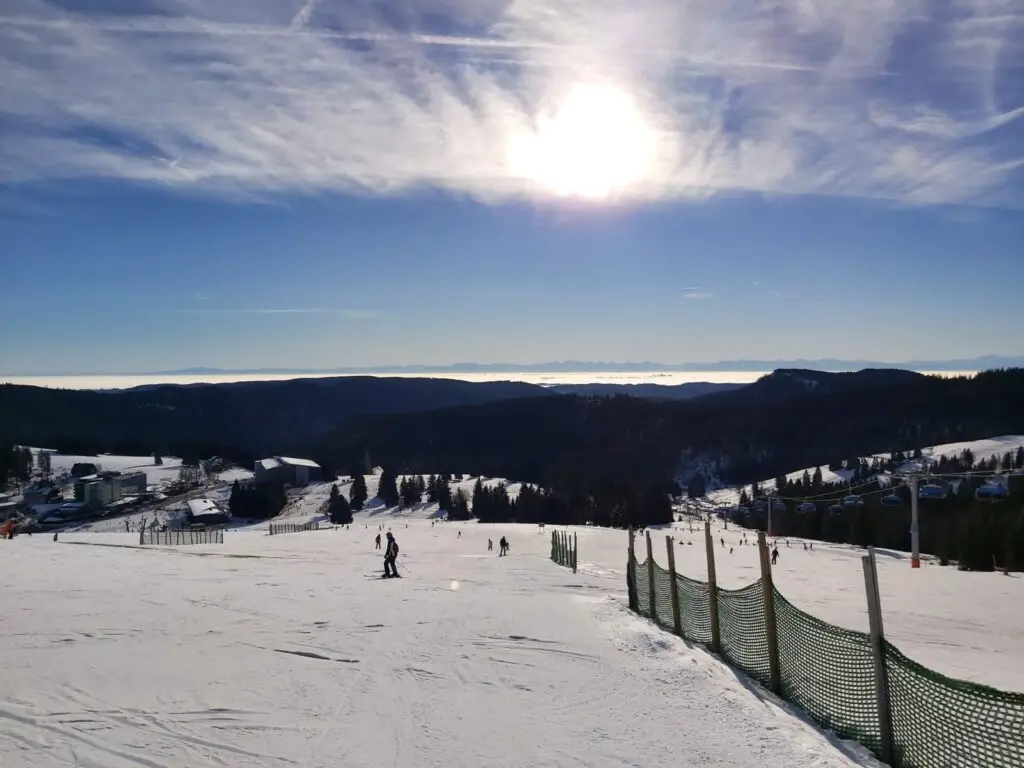 Sonnige Skipiste mit Zaun, im Hintergrund bewaldete Hügel unter blauem Himmel