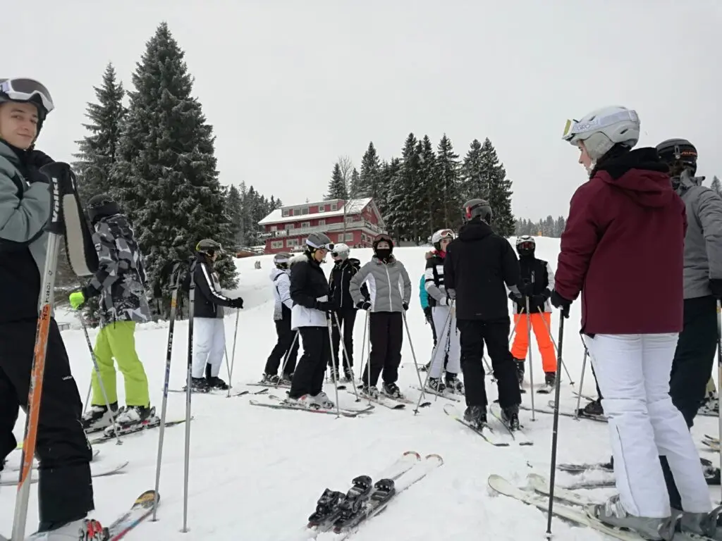 Jugendliche in Skikleidung stehen mit Ski auf einer verschneiten Fläche vor Tannen