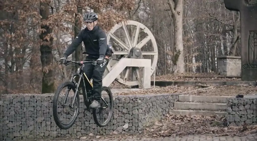 Mountainbiker mit Helm fährt auf einem Waldweg vor einem großen hölzernen Wasserrad