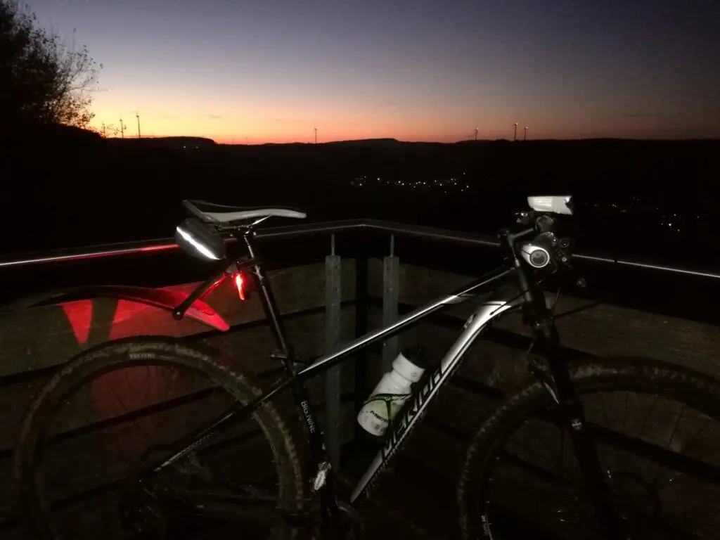Mountainbike lehnt in der Abenddämmerung an einem Geländer, dahinter ein orange leuchtender Himmel