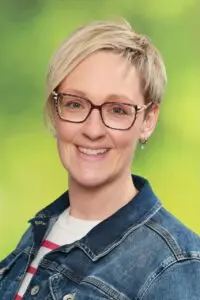 Porträt Hanna Miller, mit kurzen blonden Haaren, Brille und Jeansjacke vor grünem Hintergrund