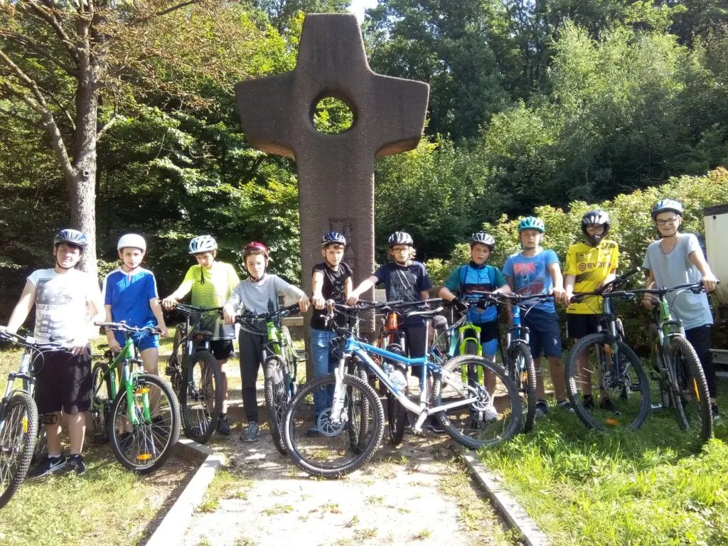 Schülergruppe mit Mountainbikes posiert vor einem großen steinernen Kreuz an einem Weg