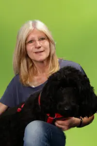 Porträt Martina Pape, mit blonden Haaren und schwarzem Hund auf dem Schoß vor grünem Hintergrund