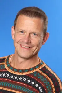 Porträt Gert Rauch, im gemusterten Strickpullover vor blauem Hintergrund