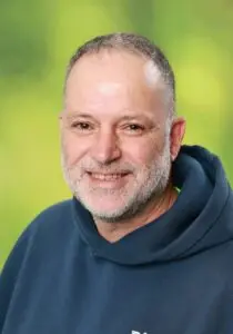 Porträt Dirk Schmeer, mit Bart im blauen Kapuzenpullover vor grünem Hintergrund