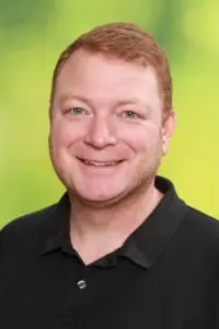 Porträt Hans Christian Schuh, lächelnd im schwarzen Poloshirt vor grünem Hintergrund