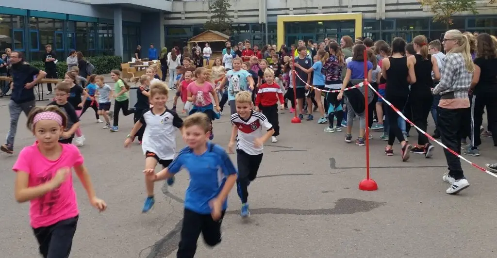Kinder laufen auf dem Schulhof an einem Absperrband entlang, Zuschauer am Rand