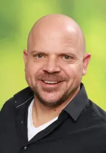 Porträt Michael Sticher, lächelnd im schwarzen Hemd vor grünem Hintergrund