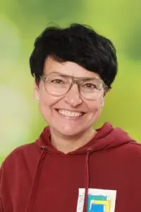 Porträt Nicole Thies, mit kurzen dunklen Haaren und Brille im roten Pullover vor grünem Hintergrund
